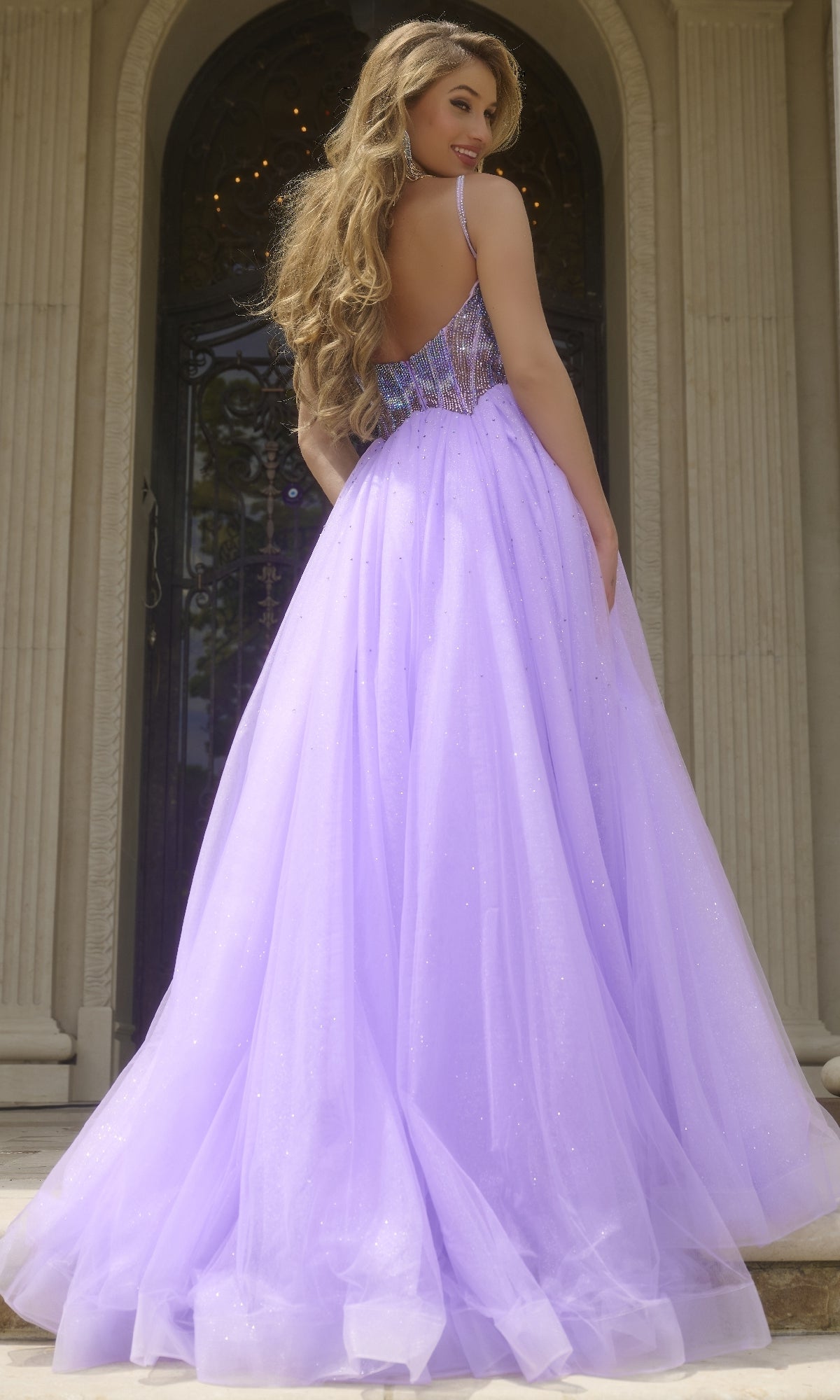 A woman in the Jovani 44511 Beaded-Corset Long Prom Ball Gown stands on steps before a grand ornate door, glancing over her shoulder.